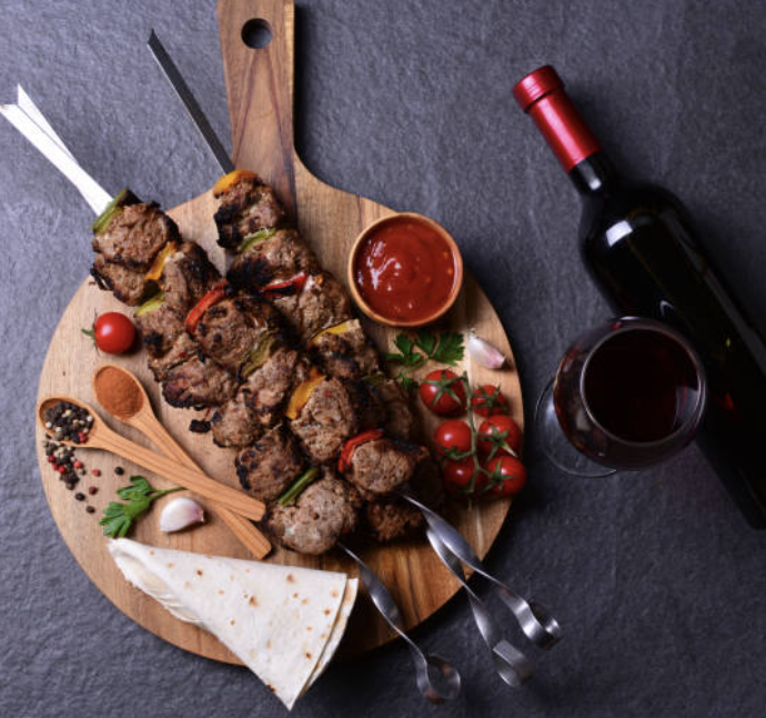 Planche en bois avec des brochettes de boeuf dessus représentant la Bourgogne, deux cuillères en bois avec des épices à l'intérieur, des tomates cerises et une bouteille de vin rouge représentant la bourgogne. 